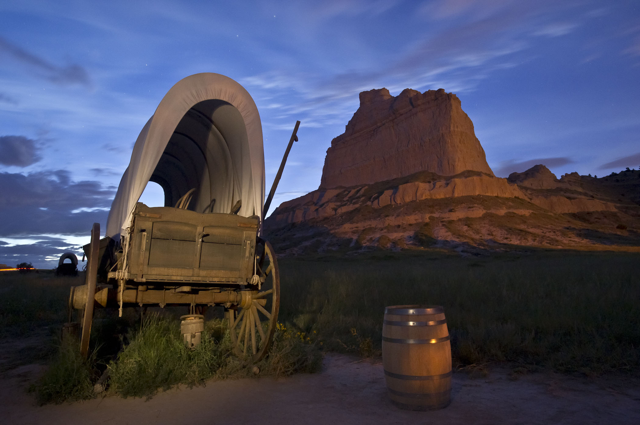 A covered wagon and barrel with Scotts Bluff National Monument towering behind it at dusk.