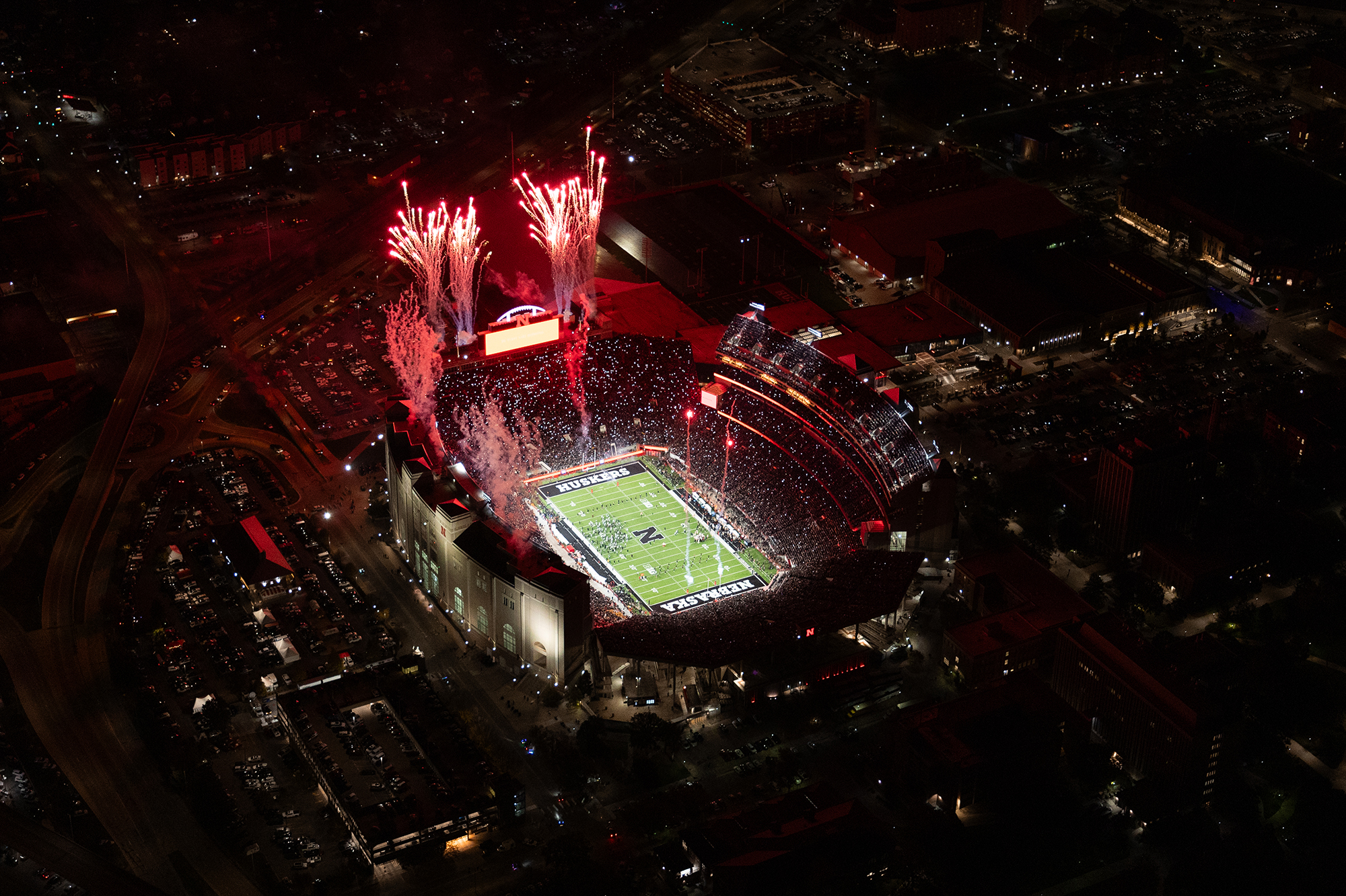 A sold out Memorial Stadium at night with red fireworks overhead during a home Nebraska Huskers football game.