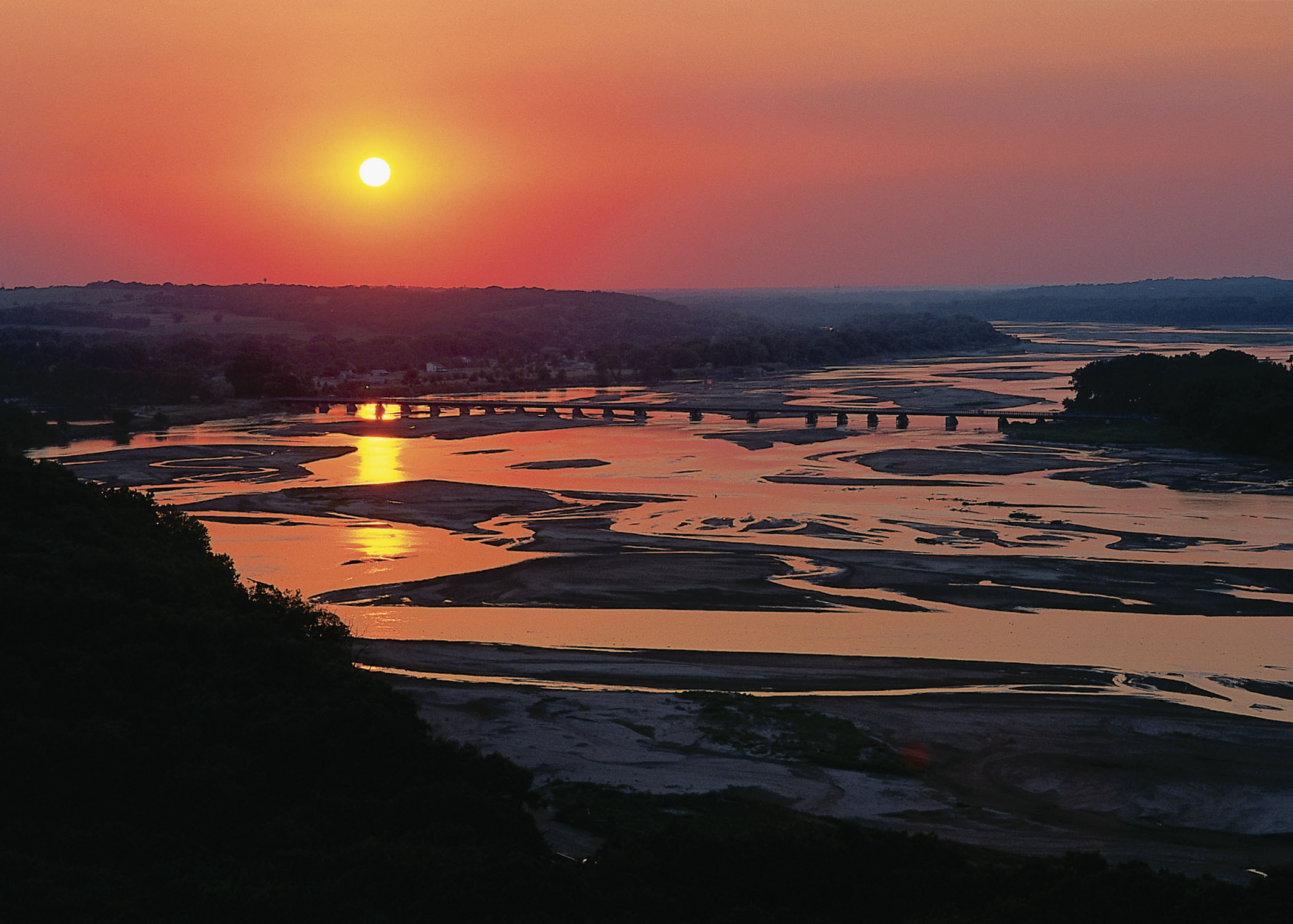 Platte River State Park with the pink and yellow sunset reflecting off the water, with a bridge running across it.