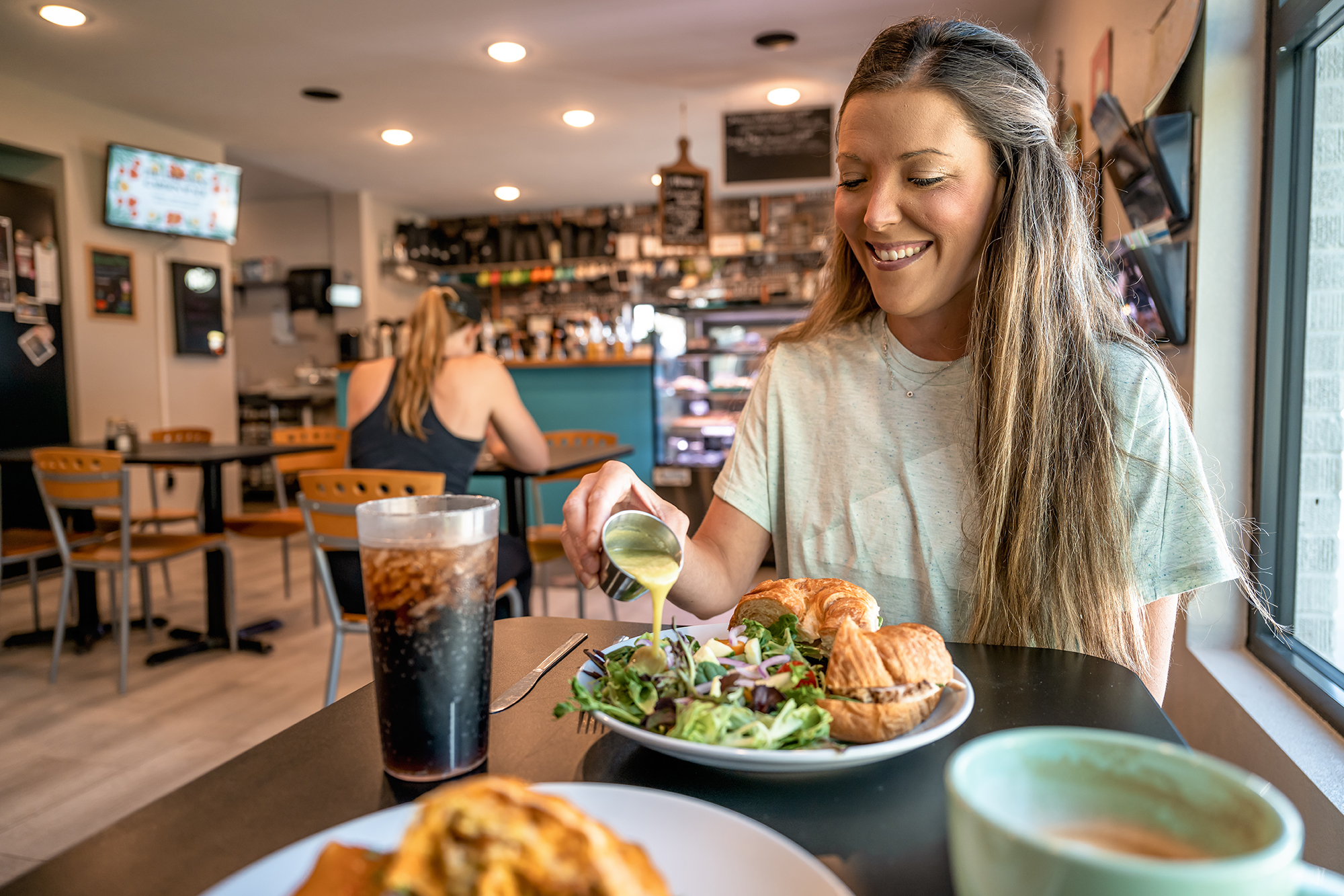 A woman pouring dressing on a salad next to a sandwich at The Mixing Bowl.