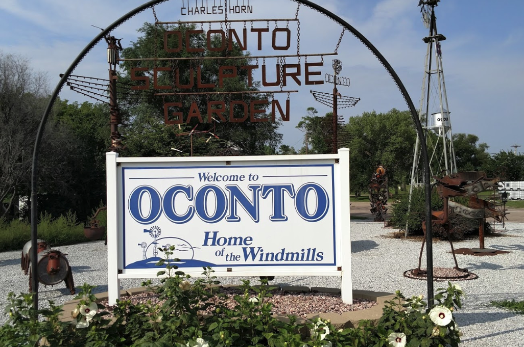 A white and blue "Welcome to Oconto" sign in front of the Charles Horn Sculpture Garden in Oconto.